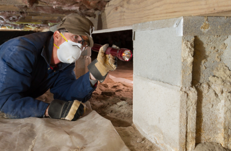 A man inspects a crawl space using a flashlight while wearing gloves and a protective mask.