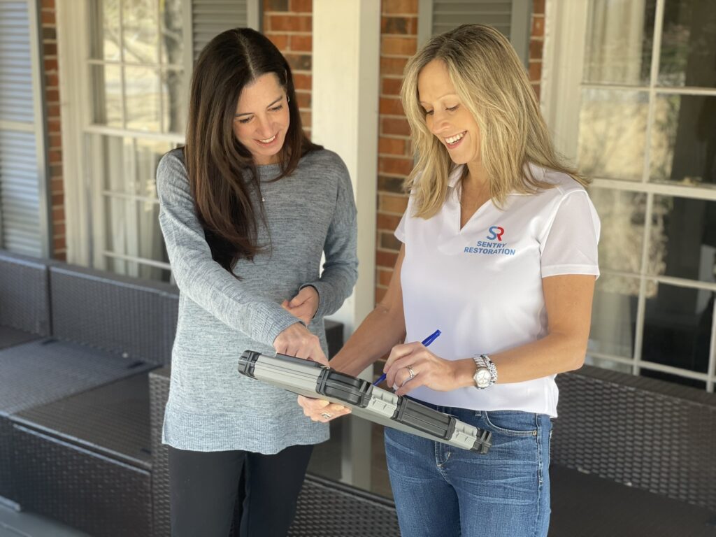 Two women discuss a tool on a porch, one pointing while the other writes notes.